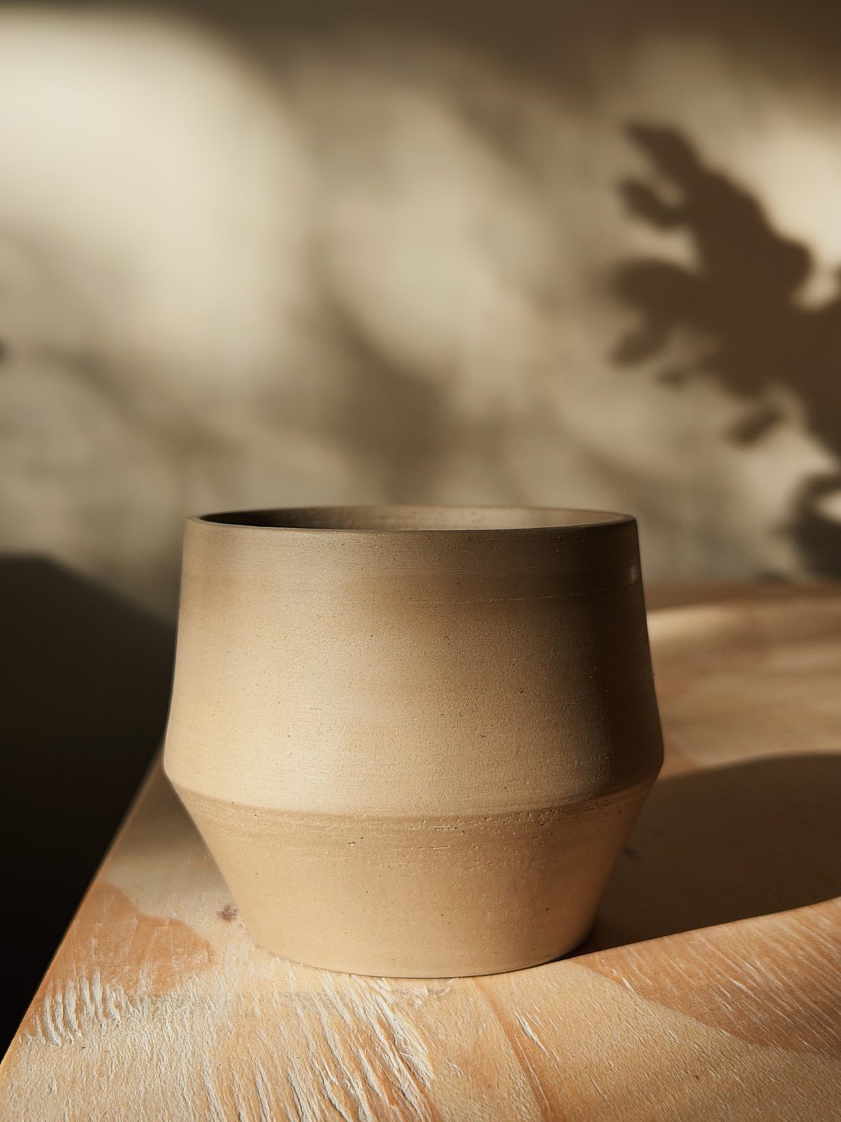 Ongeglazuurd kopje op een houten plank met schaduwen van bladeren